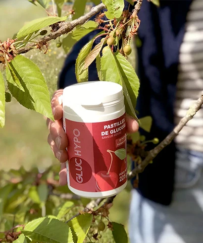 Pot de pastilles de glucose Gluc Hypo saveur cerise tenu en main dans un cerisier